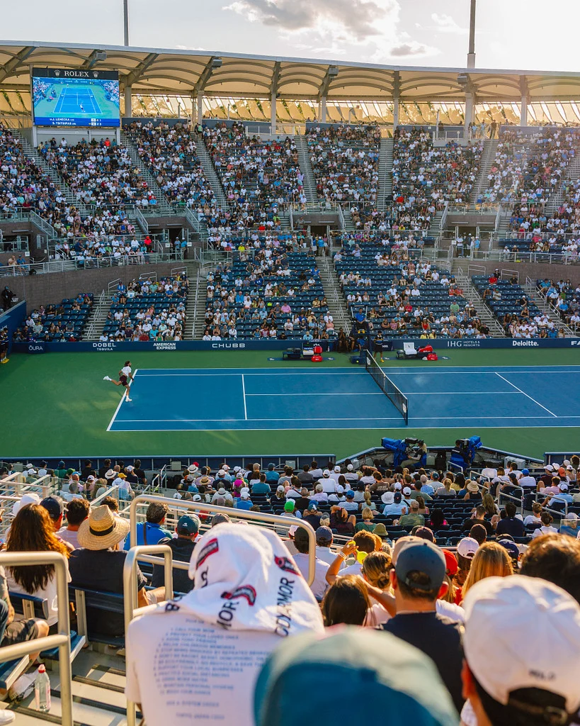 Spectators watching a tennis match on a blue court in late-afternoon sunlight